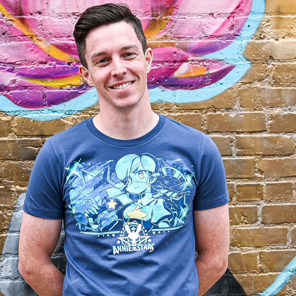 Closeup photo of a man wearing the Skullgirls Annie Of The Stars tee. The background is a wall with a nice painting of flowers that is nicely blurred.